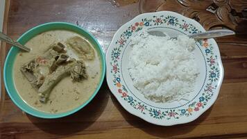 A bowl of white rice next to a bowl of soup with a spoon, served on a wooden table. photo