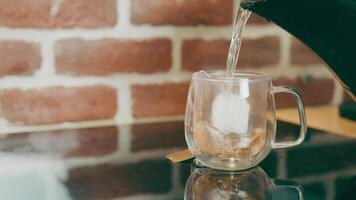 Making tea in a bag in a transparent glass cup on the kitchen table on a background of red brick wall. Tea in a bag is filled with hot water from the kettle. video