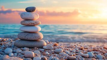 A balanced stack of smooth, grey and white stones sits on a sandy beach with the ocean and a colorful sunset in the background. photo