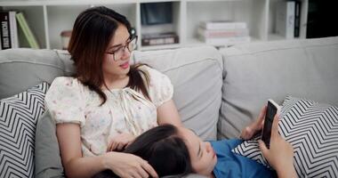 Handheld high angle view shot, A young woman is lying on her sister's lap while watching something together on her mobile phone on sofa in the living room, enjoying fun time together. video