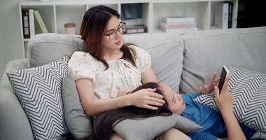 Handheld high angle view shot, A young woman is lying on her sister's lap while watching something together on her mobile phone on sofa in the living room, enjoying fun time together. video