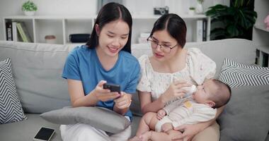 Handheld high angle view shot, Two happy women are enjoying watching something together on mobile phone while bottle-feed the baby boy on sofa in the living room. Happy family time video