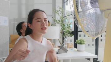 Two Asian workers are unhappy working in hot temperatures at desk in small business office in summer, uncomfortable with no air conditioner, blowing wind with fans, drinking water, and sweating. video