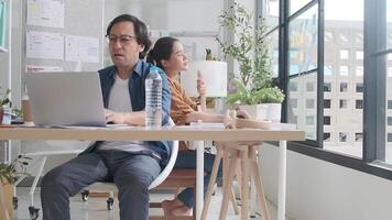 Two Asian workers are unhappy working in hot temperatures at desk in small business office in summer, uncomfortable with no air conditioner, blowing wind with fans, drinking water, and sweating. video