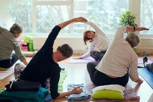 A group of senior women engage in various yoga exercises, including neck, back, and leg stretches, under the guidance of a trainer in a sunlit space, promoting well-being and harmony photo