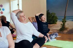 A group of senior women engage in various yoga exercises, including neck, back, and leg stretches, under the guidance of a trainer in a sunlit space, promoting well-being and harmony photo