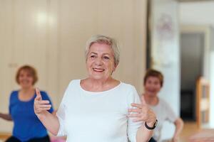 A group of senior women engage in various yoga exercises, including neck, back, and leg stretches, under the guidance of a trainer in a sunlit space, promoting well-being and harmony photo