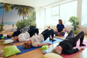 A group of senior women engage in various yoga exercises, including neck, back, and leg stretches, under the guidance of a trainer in a sunlit space, promoting well-being and harmony photo