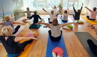 A group of senior women engage in various yoga exercises, including neck, back, and leg stretches, under the guidance of a trainer in a sunlit space, promoting well-being and harmony photo