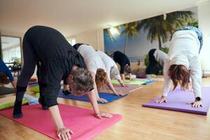 A group of senior women engage in various yoga exercises, including neck, back, and leg stretches, under the guidance of a trainer in a sunlit space, promoting well-being and harmony photo