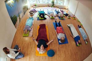 A group of senior women engage in various yoga exercises, including neck, back, and leg stretches, under the guidance of a trainer in a sunlit space, promoting well-being and harmony photo
