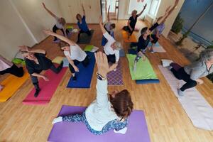 A group of senior women engage in various yoga exercises, including neck, back, and leg stretches, under the guidance of a trainer in a sunlit space, promoting well-being and harmony photo