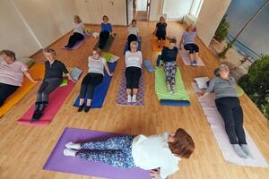 A group of senior women engage in various yoga exercises, including neck, back, and leg stretches, under the guidance of a trainer in a sunlit space, promoting well-being and harmony photo