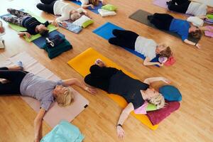 A group of senior women engage in various yoga exercises, including neck, back, and leg stretches, under the guidance of a trainer in a sunlit space, promoting well-being and harmony photo
