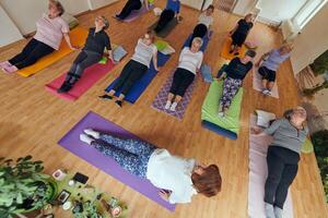 A group of senior women engage in various yoga exercises, including neck, back, and leg stretches, under the guidance of a trainer in a sunlit space, promoting well-being and harmony photo