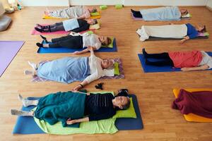A group of senior women engage in various yoga exercises, including neck, back, and leg stretches, under the guidance of a trainer in a sunlit space, promoting well-being and harmony photo