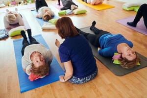 A group of senior women engage in various yoga exercises, including neck, back, and leg stretches, under the guidance of a trainer in a sunlit space, promoting well-being and harmony photo