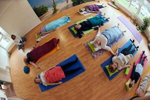 A group of senior women engage in various yoga exercises, including neck, back, and leg stretches, under the guidance of a trainer in a sunlit space, promoting well-being and harmony photo