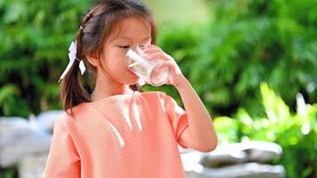 Cute Asian kid girl Drinking pure water from a glass in the garden at home. Thirsti, drink water for good health, hot weather, reduce body temperature. video