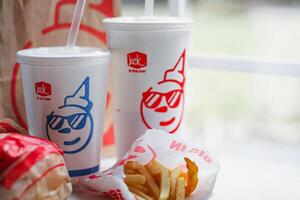 WASHINGTON, USA - August 08, 2024 - A classic fast food spread from Jack in the Box two cups of soda, a bag of fries and a wrapped burger. The cups feature the chain's iconic mascot with sunglasses. photo