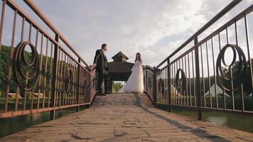 Wedding couple on a rustic bridge with gazebo in the background. A newlywed couple stands on a rustic bridge, exchanging glances while surrounded by natural greenery. video
