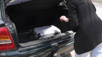 Young brunette woman carefully takes out a sleek silver carry-on suitcase from the trunk of a car on a bright sunny day, looking forward to traveling during summer holidays, video