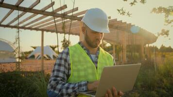 Thoughtful Caucasian male man experienced worker inspector helmet hardhat guy engineer technician typing laptop computer entering data internet pc analyzing project working blueprint construction park photo