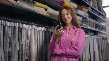 Caucasian woman tailor with mobile phone in fabric warehouse drapery shop using smartphone order home textile organic eco materials female owner small business smile choose tissues in clothing store photo