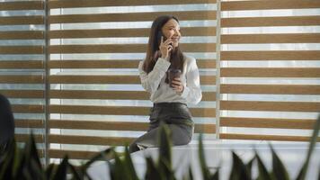 contento sonriente riendo caucásico mujer niña negocio dama mujer de negocios hablando móvil teléfono llamada simpático conversacion sentado en mesa hablar teléfono inteligente Bebiendo café mirando a ventana en oficina foto