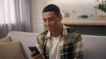 Smiling joyful cheerful African American ethnic man biracial carefree guy smile male reading message smartphone app mobile phone application device gadget browsing at home couch sofa chatting happy photo