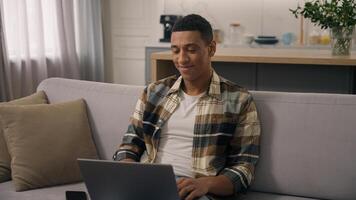 Happy smiling African American ethnic man freelancer working at home distant online work at sofa smile positive student guy studying remote with laptop using computer e-learning at couch male using pc photo
