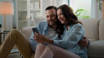 milenario Pareja caucásico hombre mujer casado marido esposa a hogar hablando utilizando teléfono inteligente juntos contento familia discutir ofertas elegir compras en línea con móvil teléfono novio demostración pulgar arriba foto
