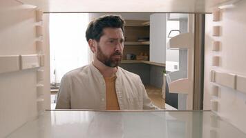 Hungry Caucasian bachelor man looking for food in empty fridge finding no meal products feeling sad disappointed poor unemployment need delivery service POV point of view from inside refrigerator photo