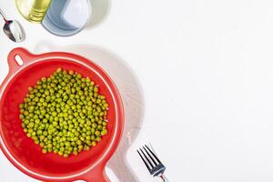 Canned peas in a red plastic colander next to an open tin can, fork and spoon photo