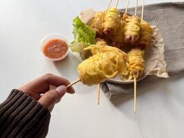 A hand holding sosis mie gulung. Rolled noodle sausages are made from sausages that are skewed then rolled in dry noodles and then fried with beaten egg. Served on a plate with sauce photo