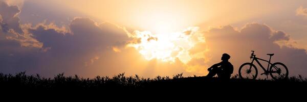 Silhouette of Person Sitting Beside Bicycle at Sunset with Dramatic Sky and Clouds in Background photo