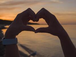 Hands form a heart-shaped silhouette against a sunset, where the suns glow softly illuminates the heart-shaped outline. This heart-shaped gesture resonates with warmth and love as day turns to night. photo
