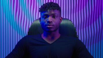 Portrait African American gamer guy programmer hacker man male user sit in black chair relaxing in neon futuristic room studio violet blue futurism innovation computer technology data internet cyber video