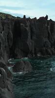 Top view of black rocks with blue waves on cloudy day. Clip. Black stone cliffs on ocean shore on cloudy day. Northern coasts on cloudy summer days video