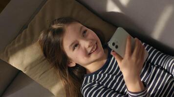 Caucasian European little girl lying on sofa holding using mobile phone smartphone child gadget online texting sms social networks smiling laughing enjoyment watching funny internet chatting kid video