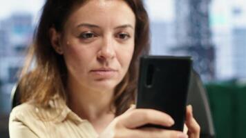 Female CEO taking a break from work and answering text messages on her smartphone, discussing with partners or friends in her high end business office. Social media updates online. Camera A. video
