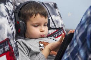 A boy sitting on a sofa in the room and listening to music or an e-learning class on his tablet computer with headphones. A child watching movie on digital tablet with headphones photo