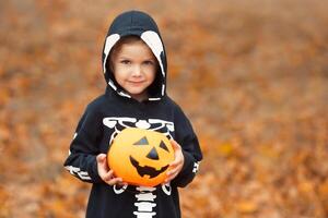 Close-up portrait of a child boy 4-5 years old in a skeleton costume holding a Halloween candy bucket on a background of autumn leaves. Copy space. photo