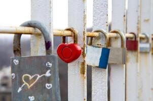 Close-up of various padlocks, including a red heart-shaped lock, attached to a rusty white metal railing symbolizing love, commitment, and memories on a bridge photo