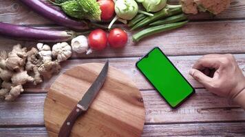 A man is holding a smartphone with a green screen on a wooden table with vegetables and a knife video