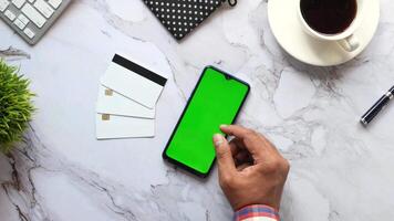 A man using a smartphone with a green screen on a desk with a cup of coffee and a video