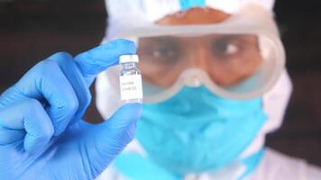 A man in a white coat and blue gloves holds up a vial of a vaccine video
