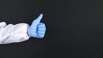 A person in a white lab coat and blue gloves is giving a thumbs up video