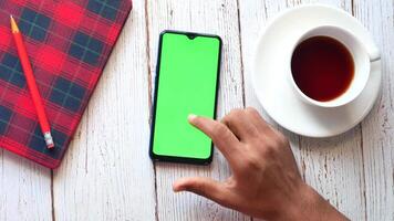 A hand holding a smartphone with a green screen and a cup of coffee on a wooden table video