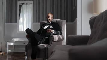 Groom Relaxing in a Chair Before the Wedding. A groom, dressed in a sharp black tuxedo, sits relaxed in a chair, basking in soft light streaming through a nearby window. video